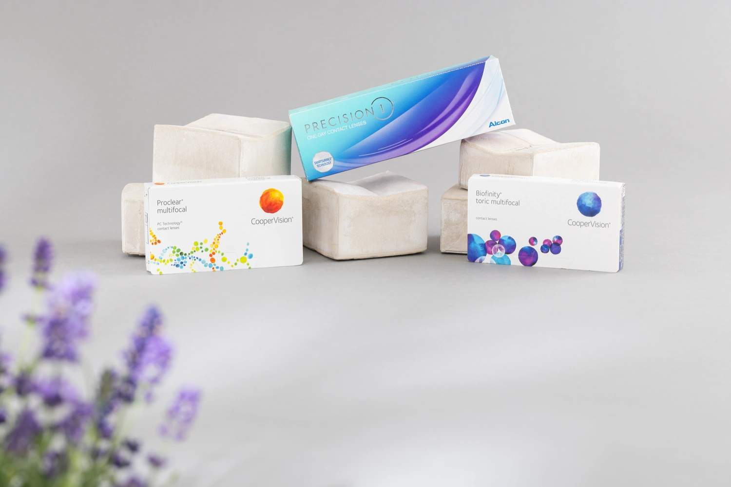 Kontaktlinsen kaufen Drei verschiedene Kontaktlinsen-Boxen arrangiert mit einer lila Blume im Vordergrund.